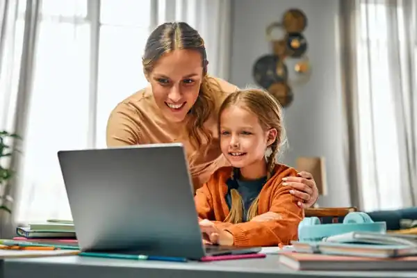 A parent leaning in to help her child with schoolwork at the kitchen table